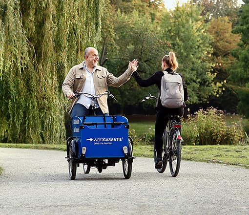 Zwei Menschen kommen sich auf dem Fahrrad entgegen und klatschen sich ab. Der Mann fährt ein WERTGARANTIE-Lastenrad.
