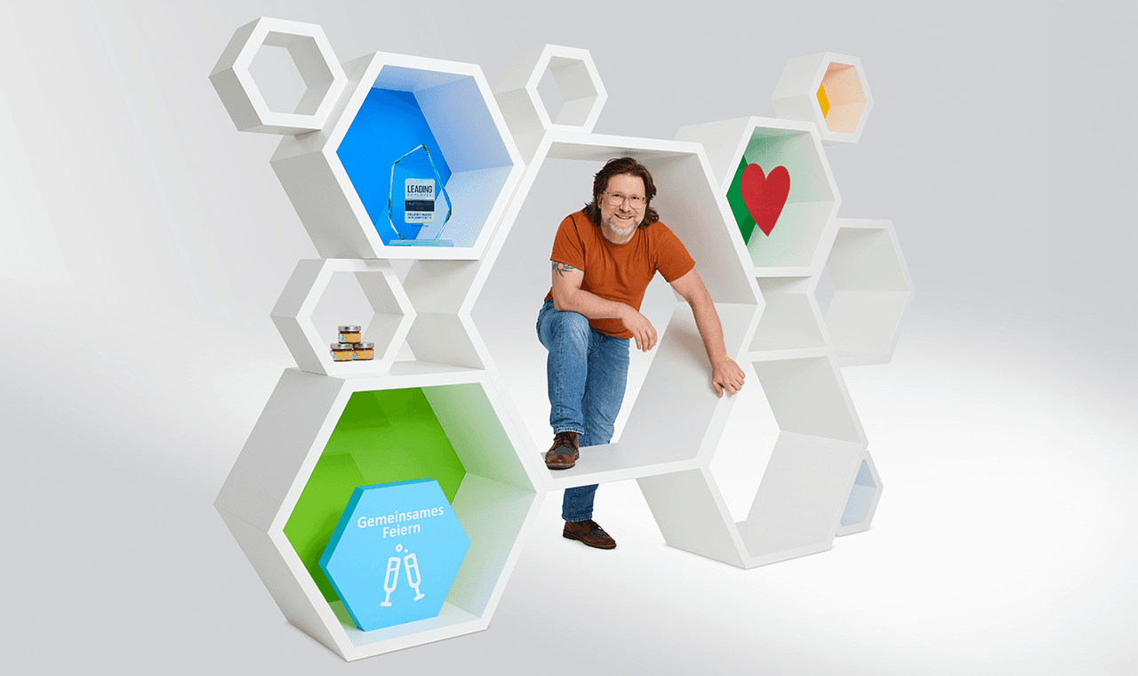 Ein Mitarbeiter steht in einem Modell aus weißen Waben gefüllt mit einem Award, Honiggläsern und einem Schild  "gemeinsames Feiern".