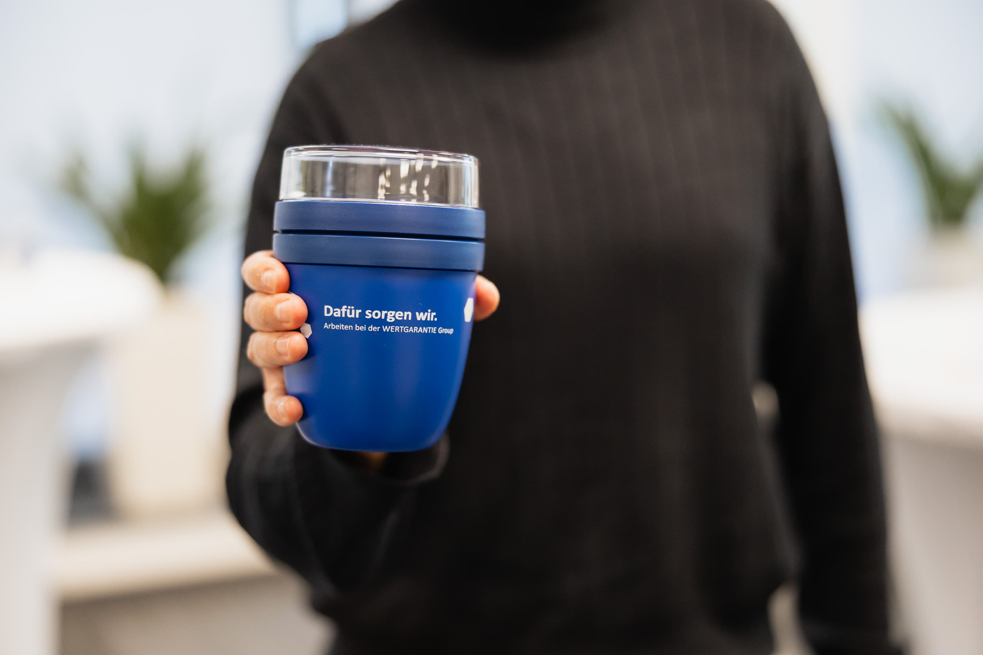 Eine Person hält einen blauen Mepal Müslibecher mit dem WERTGARANTIE Group Logo in die Kamera