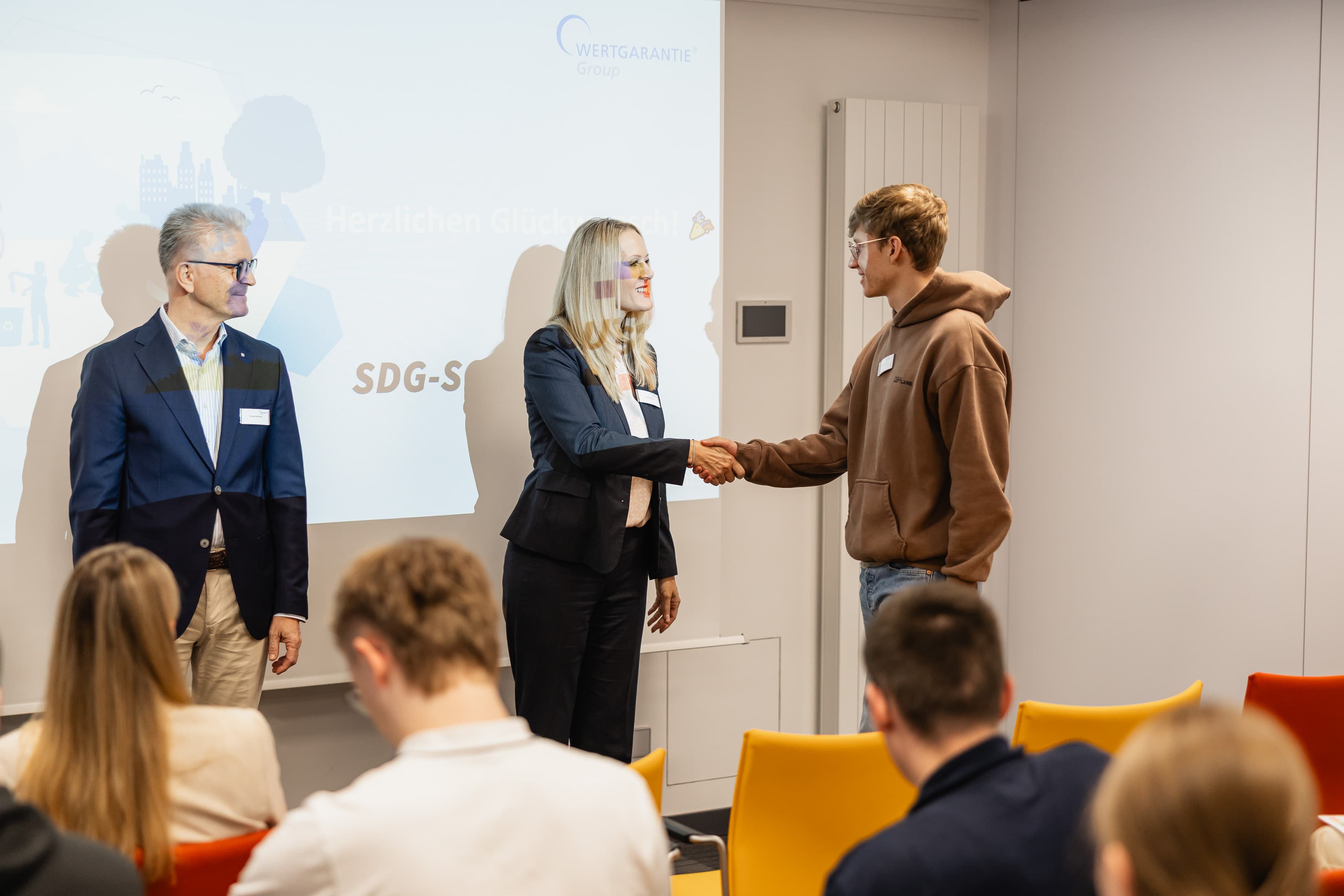 Zwei Personen stehen vor einer Präsentation, eine Frau schüttelt einem jungen Mann die Hand.
