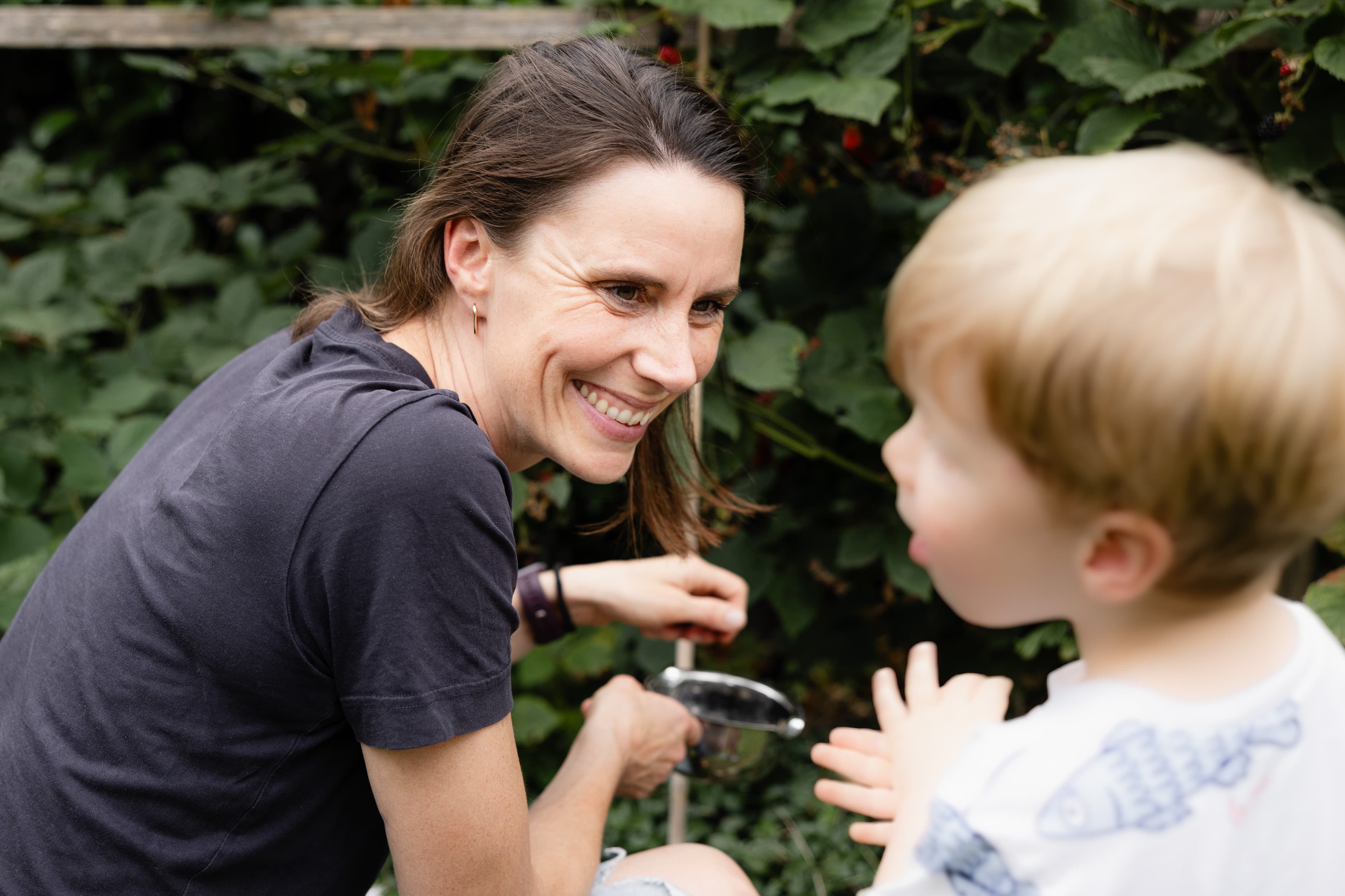 Eine Mitarbeiterin pflückt mit ihrem kleine Sohn Brombeeren im Garten. Sie lächelt ihren Sohn an, der unscharf im Vordergrund zu erkennen ist.