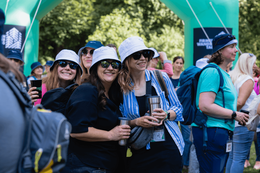 Drei Mitarbeiterinnen der WERTGARANTIE Group beim Firmenwandertag vor dem Start mit gebrandetem Anglerhut lachen in die Kamera.