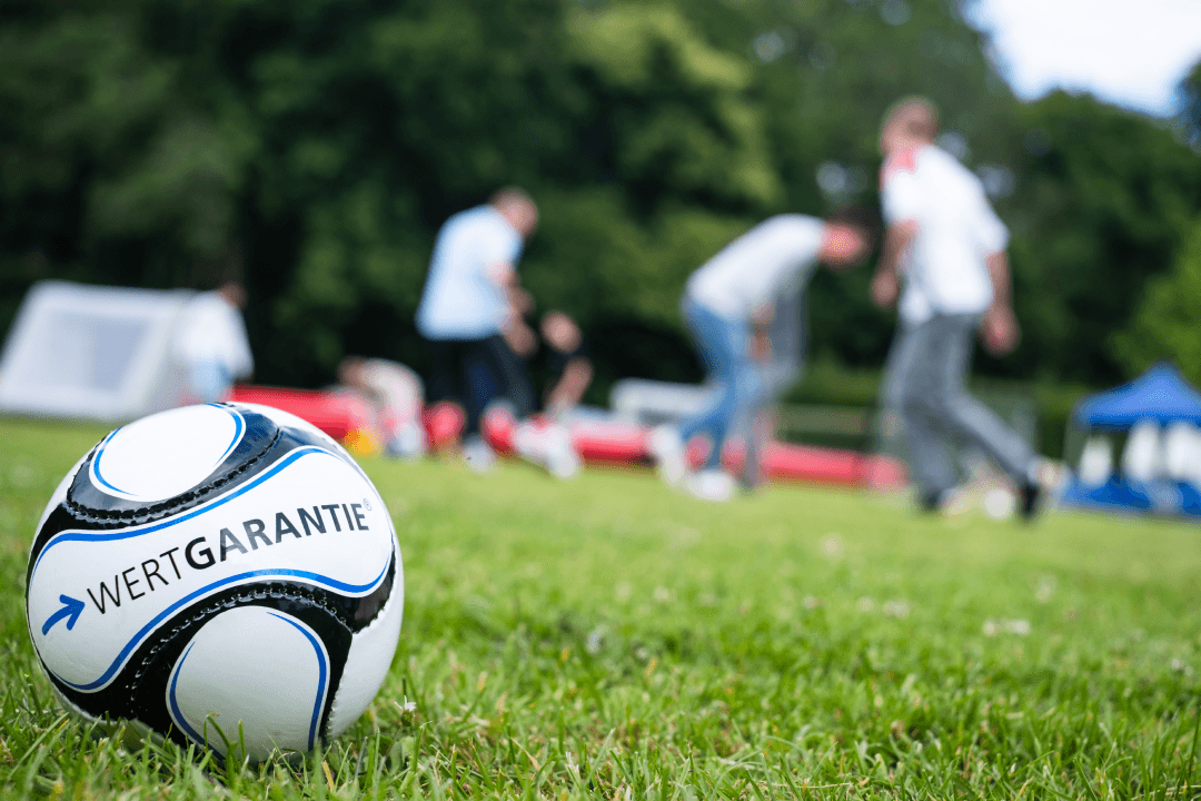 Im Vordergrund liegt ein WERTGARANTIE Fußball auf dem Rasen während im Hintergrund unscharf Mitarbeitende Fußball spielen.