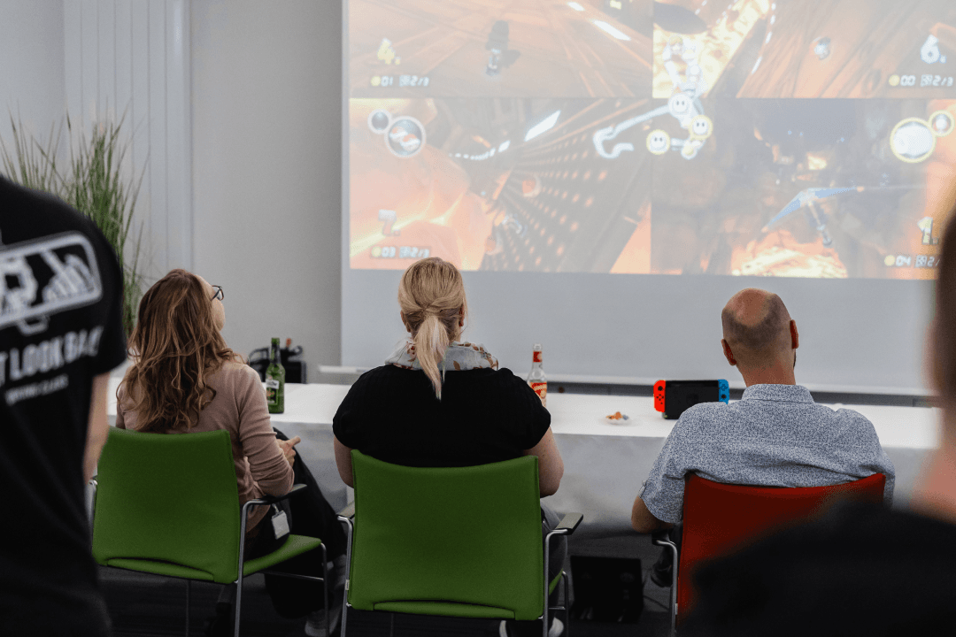 Drei Mitarbeitende der WERTGARANTIE Group sitzen nebeneinander und schauen auf eine Leinwand auf der sie Mario Kart spielen.