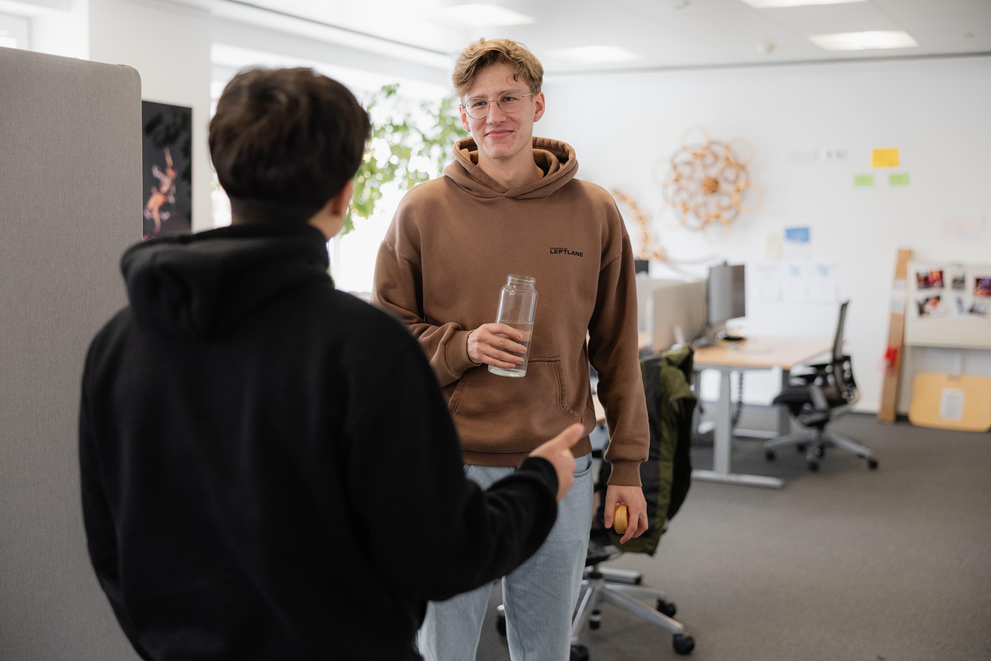 Unser Dualer Student steht im Büro, schaut in die Kamera und unterhält sich mit einem Kollegen der mit dem Rücken zur Kamera steht.