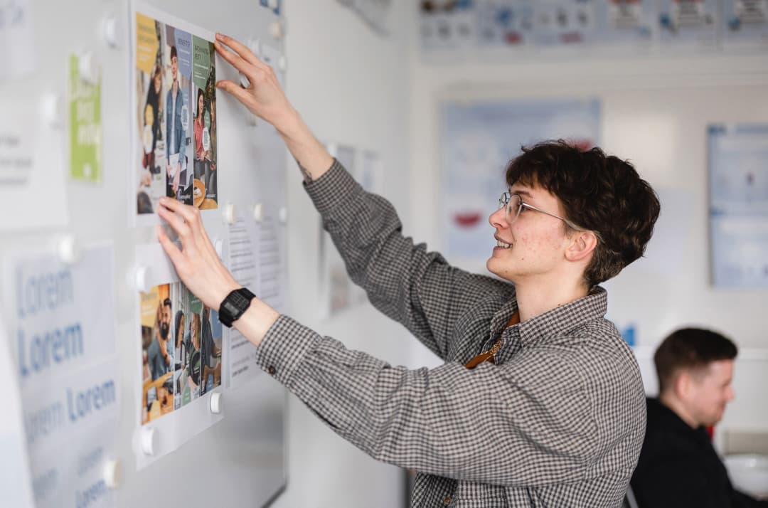 Ein Auszubildener zum Mediengestalter steht an einer Magnetwand und hängt ein Bild auf.