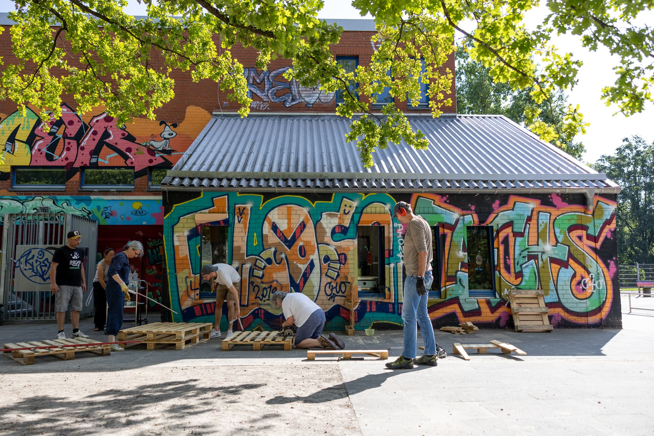 Mehrere Personen stehen vor einem graffitibesprühtem Gebäude und bauen Paletten zusammen.