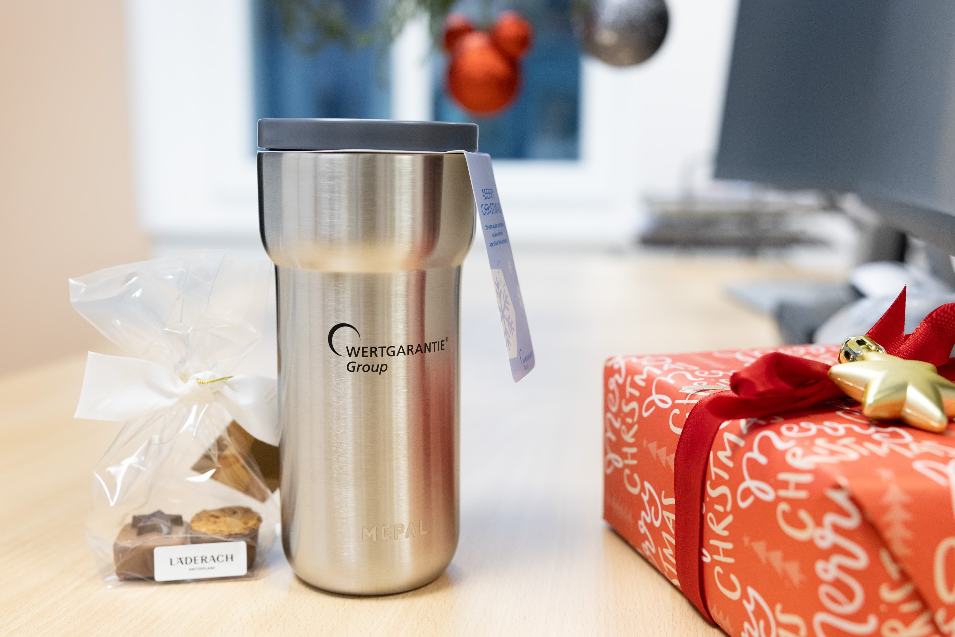Ein Mepal Thermobecher mit der Aufschrift "WERTGARANTIE Group" steht neben Pralinen und einem eingepackten Weihnachtsgeschenk.