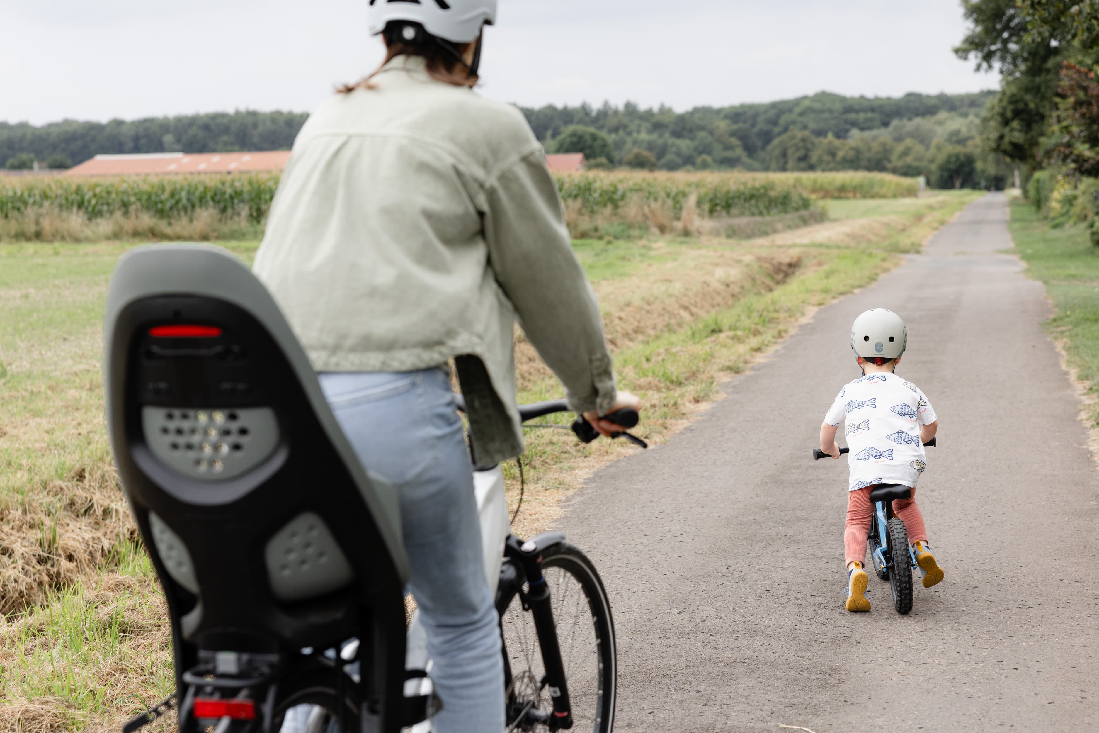 Eine Mitarbeiterin fährt mit ihrem Sohn auf dem Feldweg nebeneinander Fahrrad. Beide sind von hinten zu sehen und tragen einen Fahrradhelm.
