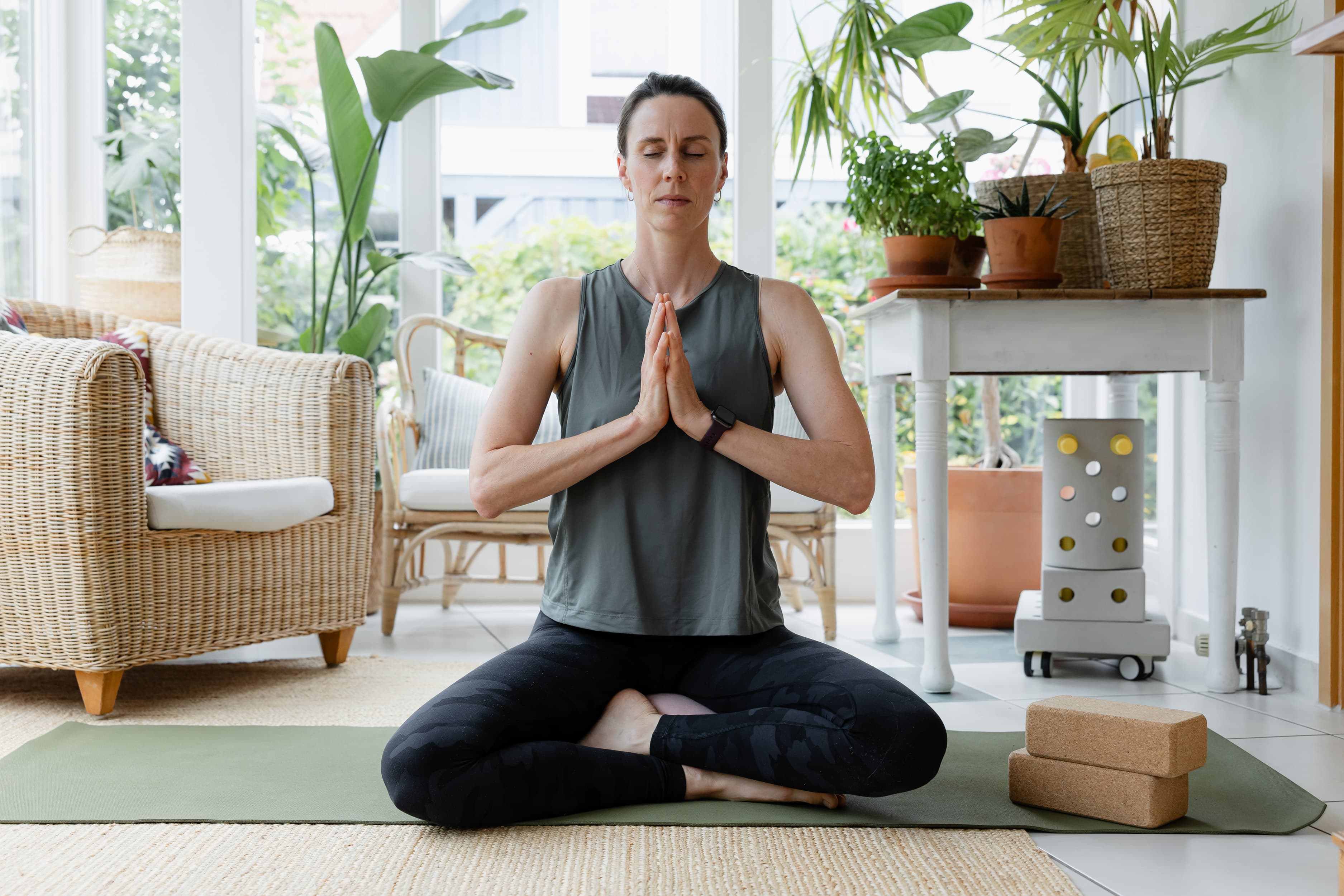 Eine Mitarbeiterin sitzt mit geschlossenen Augen im Homeoffice auf ihrer Yogamatte im Schneidersitz und führt die Hände aneinander vor ihrem Herz.