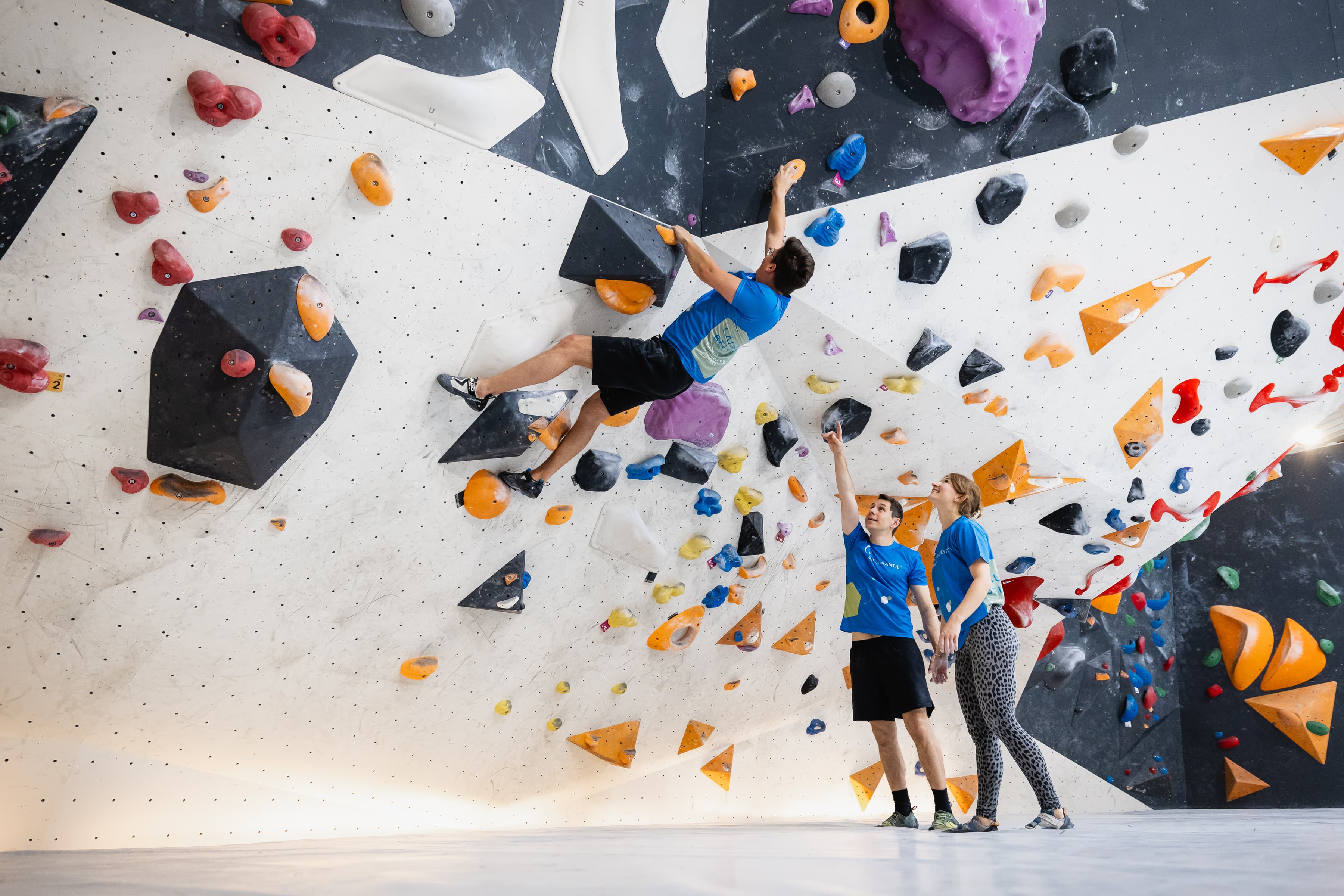 Drei Mitarbeitende in der Boulderhalle urban apes in Hannover. Ein Mitarbeiter hängt an der Kletterwand und zwei Mitarbeitende stehen auf dem Boden und zeigen auf ihn.