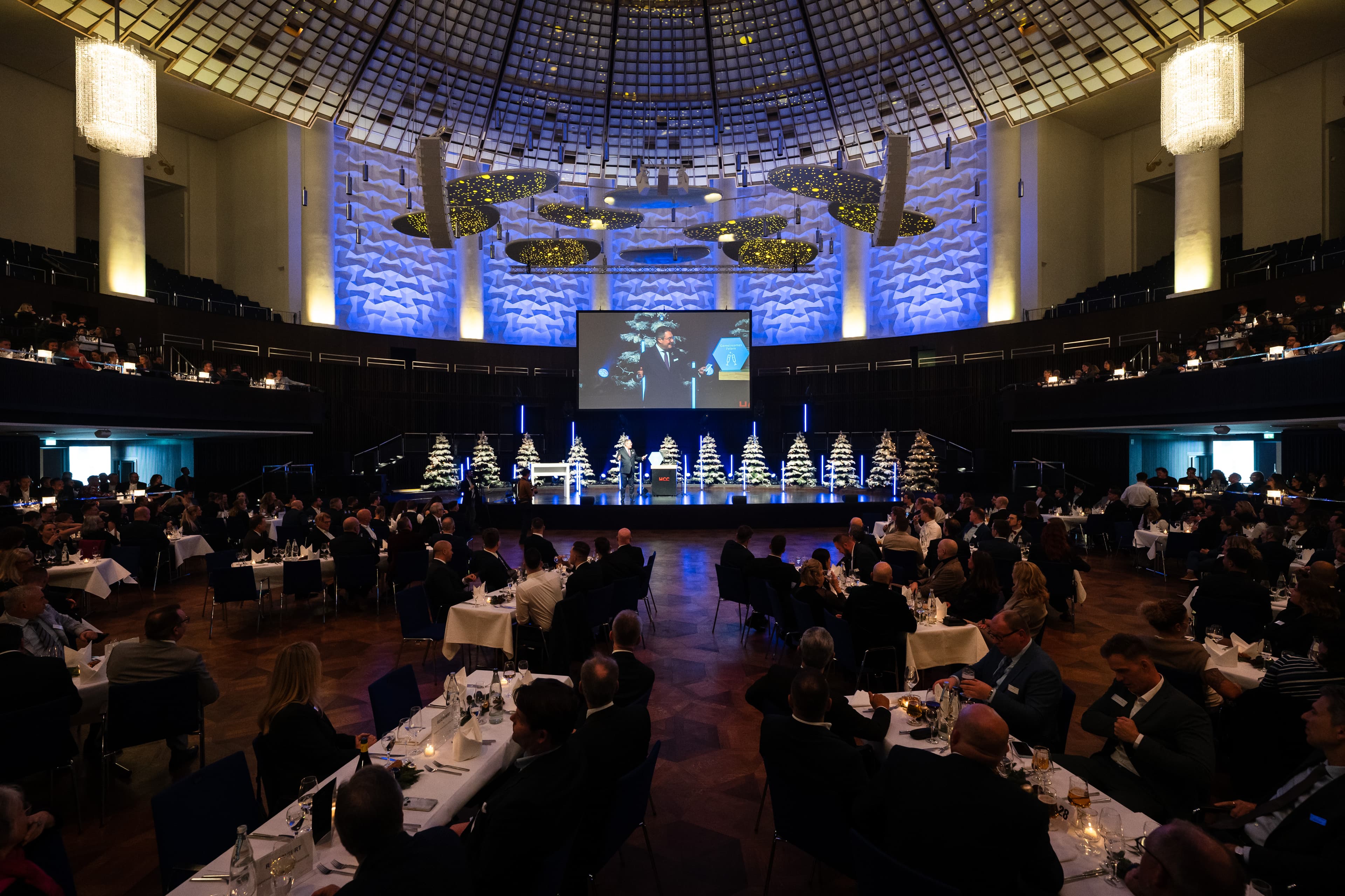 Ein Blick in den Kuppelsaal an unserer Jahresabschlussfeier. Im Vordergrund gut besuchte Tische und im Hintergrund die große Bühne auf der unserer Vorstandsvorsitzender Patrick Döring seine Rede hält.