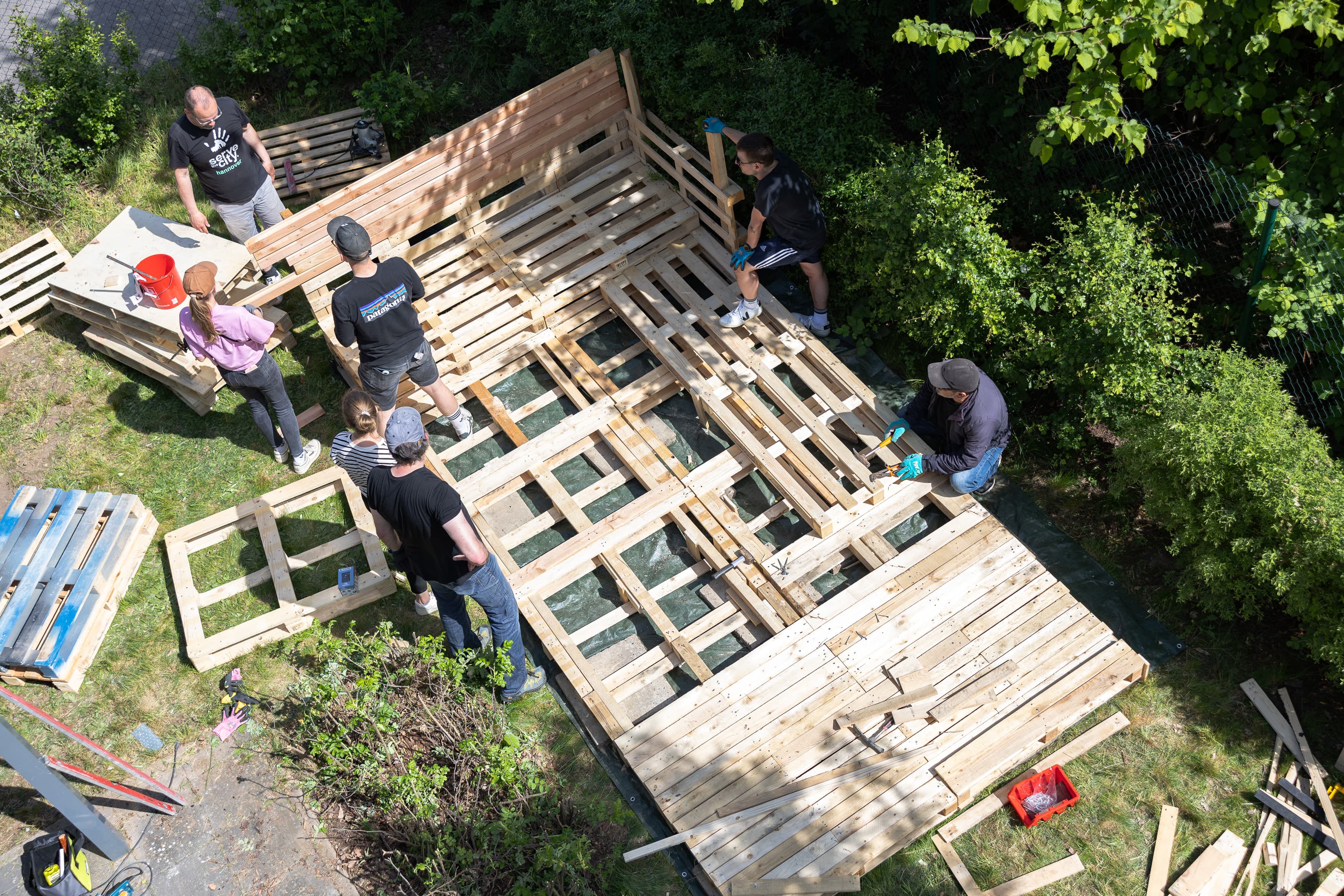 Eine Gruppe von Mitarbeitenden der WERTGARANTIE Group bauen in einer Geflüchtetenunterkunft gemeinsam Palettenmöbel.